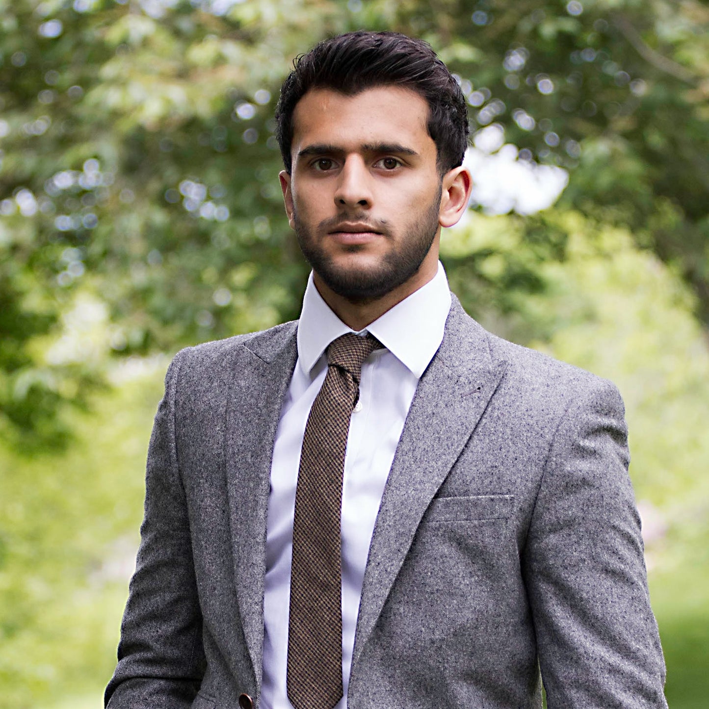 A man wearing a brown tweed necktie with a houndstooth pattern. The tie is matched with a white shirt and a grey suit made by Daisy and Oak Studio.